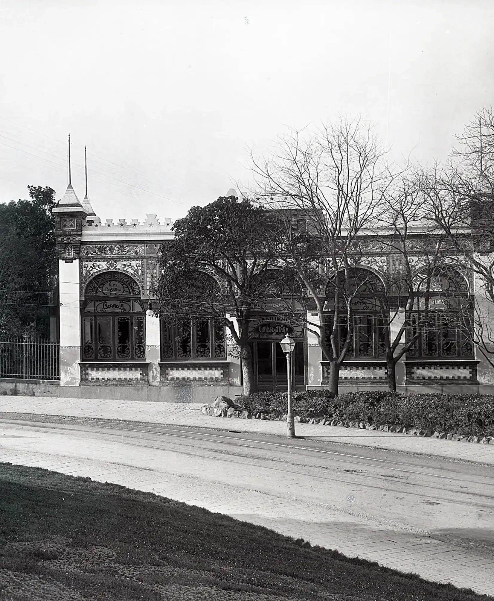 Fotografia storica in bianco e nero del Giardino d’Italia, con un elegante edificio decorato da motivi ornamentali e grandi finestre ad arco. Alberi e aiuole costeggiano la facciata, mentre in primo piano si vedono un lampione, il marciapiede e la strada.